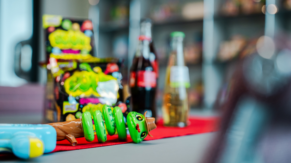 Eine bunte Spielzeugschlange liegt auf einem roten Tuch, im Hintergrund sind Snacks und Flaschen zu sehen.
