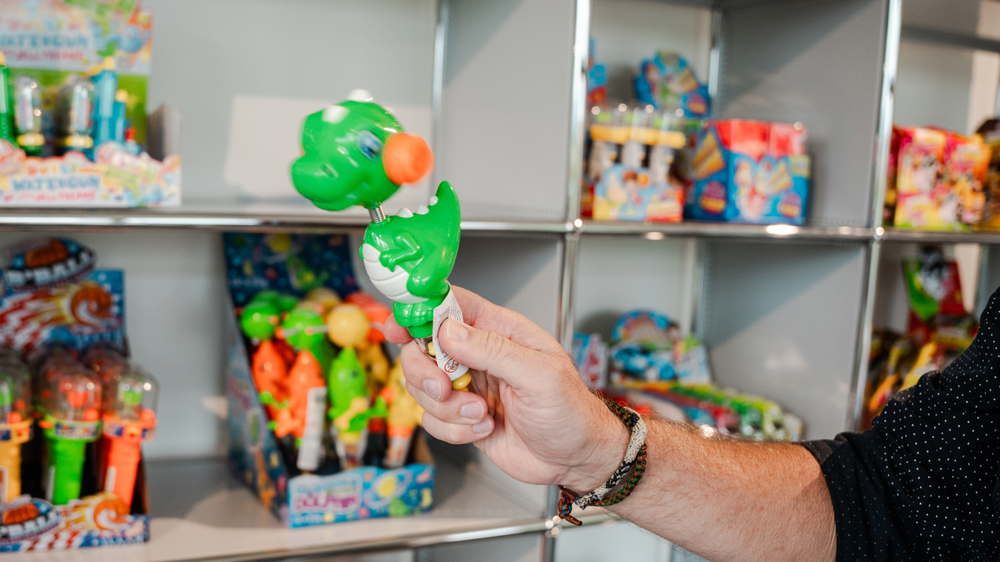 Ein gr&uuml;ner Dinosaurier-Spielzeug mit einem orangefarbenen Ball, gehalten von einer Person, vor einem Regal voller bunter Spielwaren.