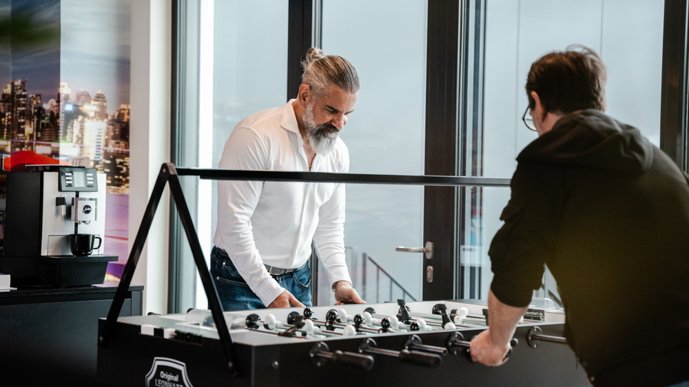 Zwei M&auml;nner spielen am Tischfu&szlig;ball in einem modernen Raum mit Fensterblick. Ein Kaffeeautomat steht daneben.