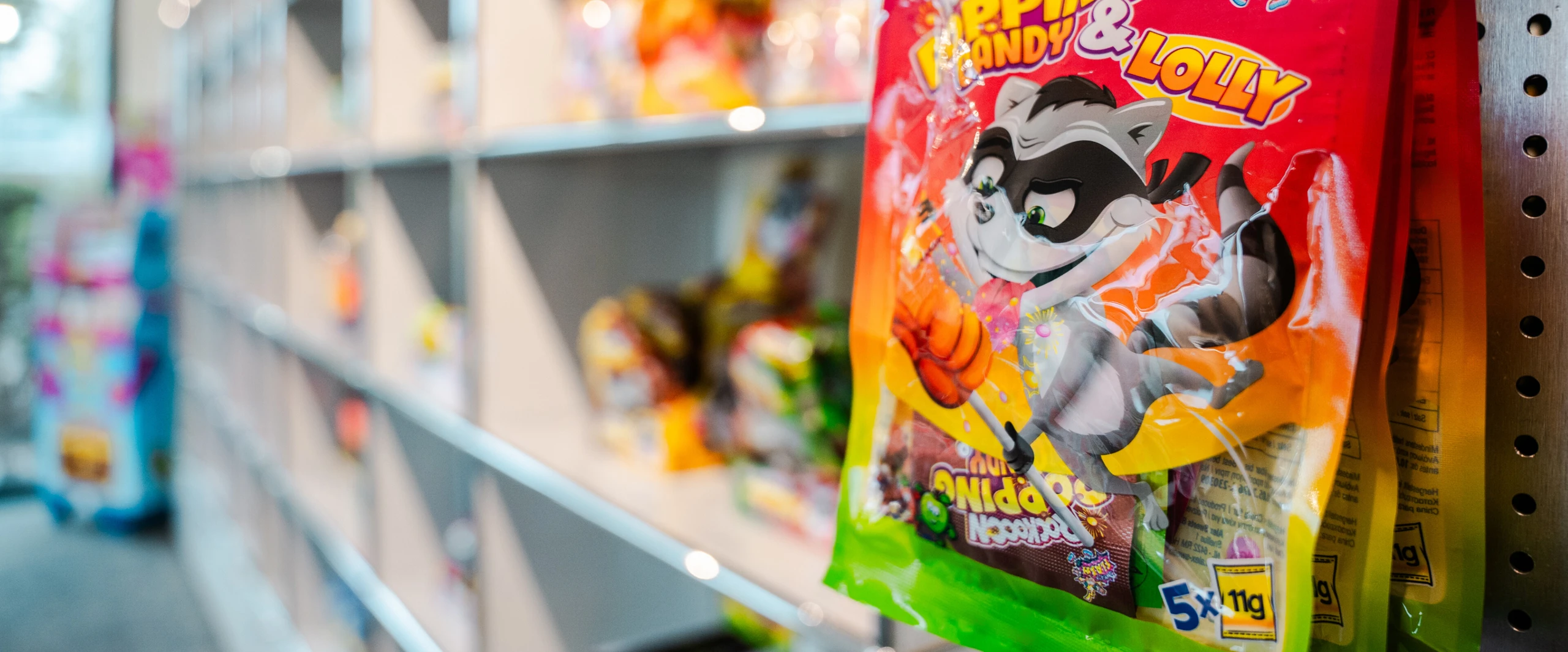 Eine Tüte mit Bonbons, die ein cartoonartiges Waschbärenmaskottchen zeigt, hängt an einem Regal mit weiteren Süßigkeiten im Hintergrund.