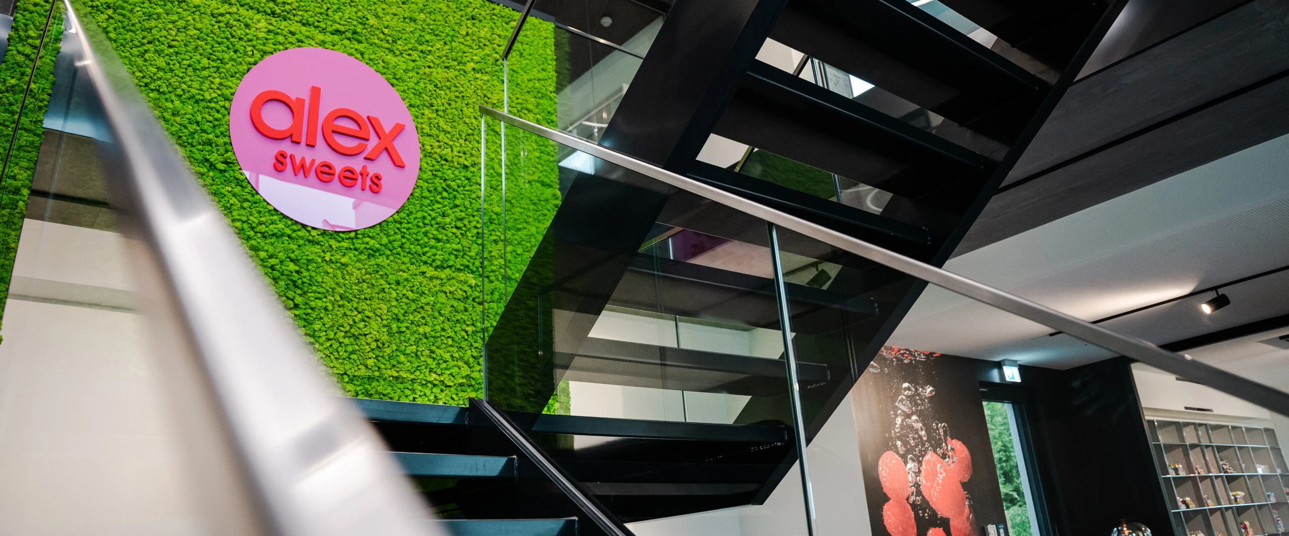 Moderne Treppe mit Glasgeländer und einer hellgrünen Mooswand. Das Logo „alex sweets“ in rosa ist sichtbar.