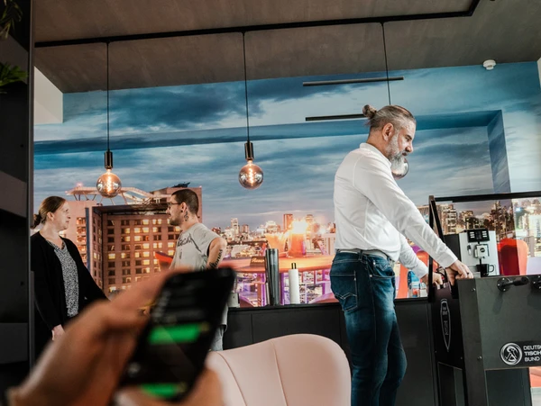 Ein Mitarbeiter spielt Tischfußball in einem modernen Büro mit Stadtlandschaft-Dekor, während andere Kollegen sich unterhalten und ein weiterer auf einem Smartphone tippt.