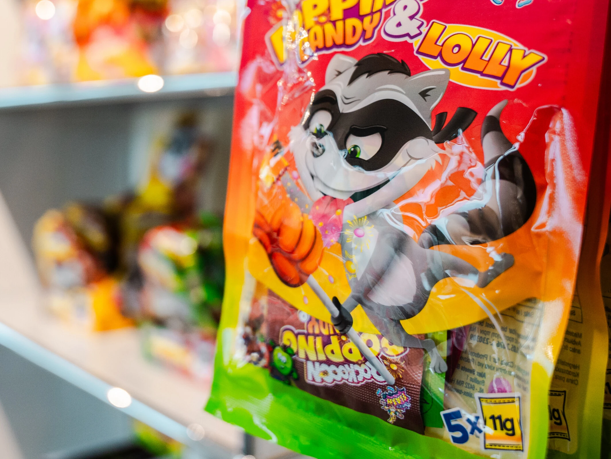 Eine Tüte mit Bonbons, die ein cartoonartiges Waschbärenmaskottchen zeigt, hängt an einem Regal mit weiteren Süßigkeiten im Hintergrund.