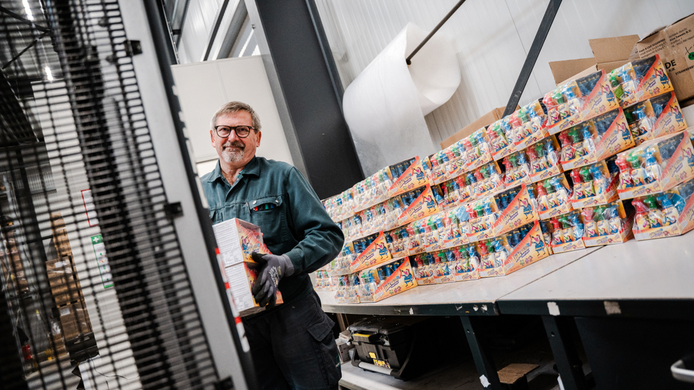 Un employ&eacute; dans un entrep&ocirc;t tient une caisse de produits, tandis qu'en arri&egrave;re-plan, on voit des piles d'emballages.
