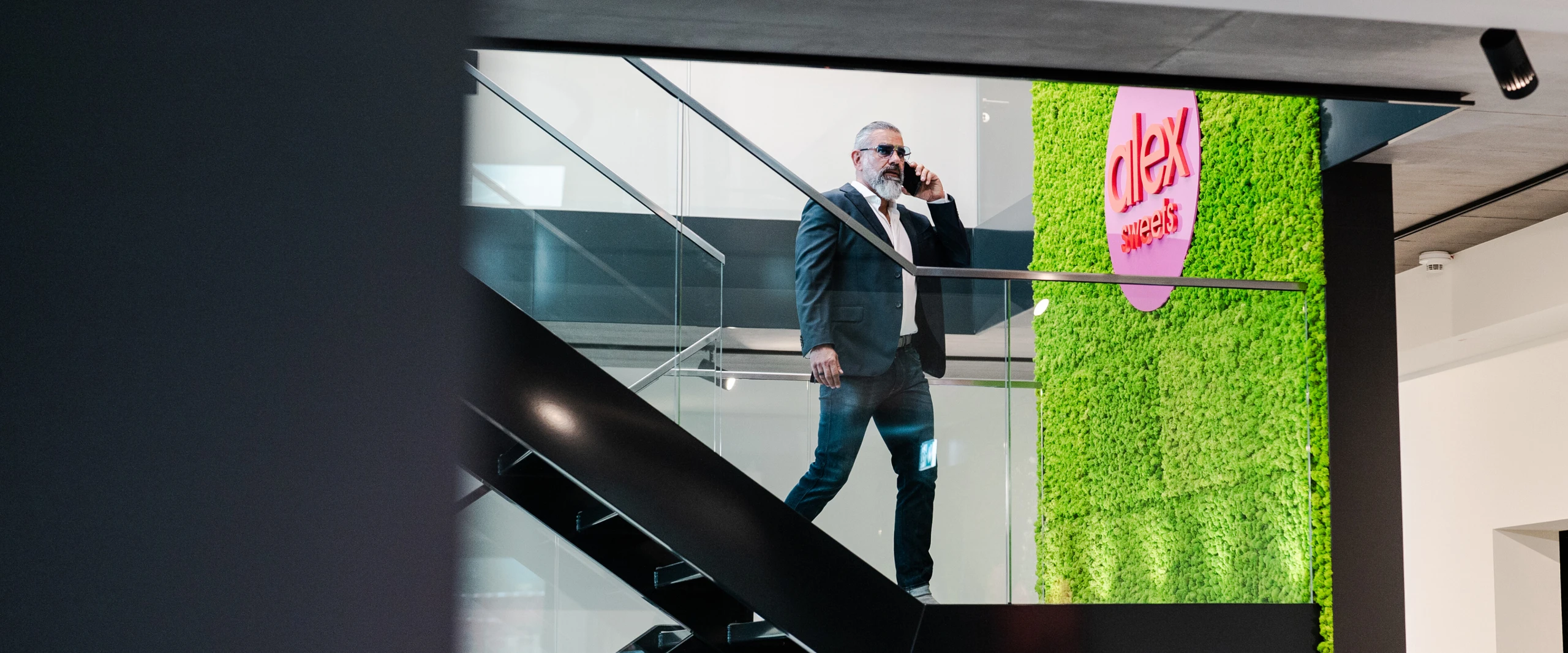 Un homme en costume et lunettes de soleil parle au téléphone et descend un escalier dans une pièce moderne avec un mur vert et l'inscription 